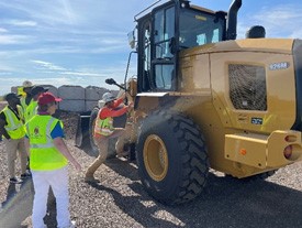 Workers at a worksite watching one person in safety gear climb onto heavy equipment.
